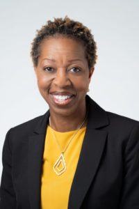 Valerie smiles, wearing a yellow shirt under a black blazer with a sliver necklace. Her photo is taken against a white, gray background.