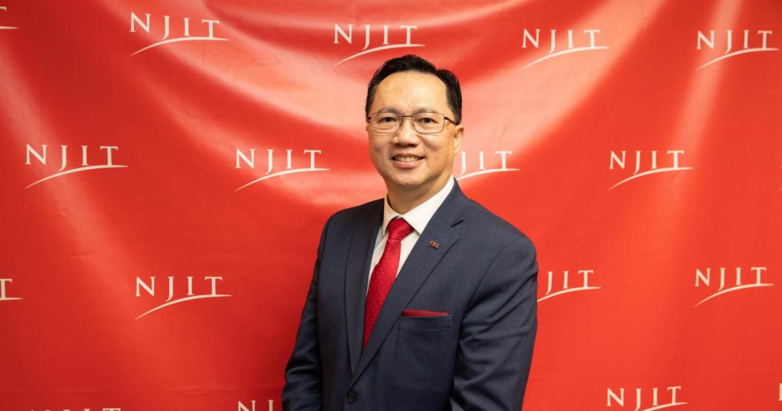 President Lim stands facing the camera in a suit and red tie in front of an "NJIT" backdrop smiling.