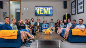 A group of 20 or so researchers of different ages sit around a U-shaped couch smiling. The name of the lab "VEMI" is on a screen in yellow and blue in the background and there are yellow snflowers in the middle of the table.