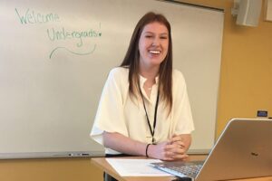 Kylee, a student at UNH with long red hair, smiles from a presentation podium behind her laptop and in front of a whiteboard